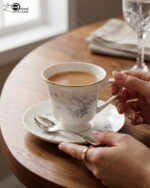 A person holding a white bone china teacup filled with milk tea, featuring a grey floral design and gold rim, resting on a matching saucer with a silver spoon on a wooden table.