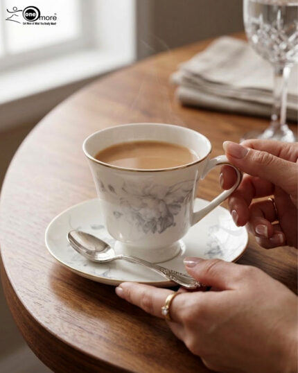 A person holding a white bone china teacup filled with milk tea, featuring a grey floral design and gold rim, resting on a matching saucer with a silver spoon on a wooden table.