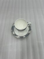 A high-angle shot of a premium super bone china cup and saucer filled with milk tea, featuring a grey floral pattern and silver spoon, set on a wooden table in a cozy living room with green sofas and a fireplace.