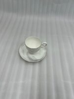 A luxury white super bone china teacup filled with creamy tea, featuring gold-rimmed edges and a delicate grey leaf pattern, resting on a matching saucer on a dark wooden table in a cozy, blurred library background