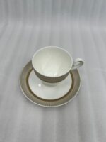 A luxury super bone china tea cup and saucer with gold pinstripe and laurel wreath patterns, filled with milk tea on a wooden table in a classic vintage living room setting with a fireplace and green armchairs