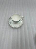 A luxury super bone china tea cup with a floral mint-green band and gold rim, filled with coffee, resting on a matching saucer on a polished wooden table in a classic living room with a fireplace
