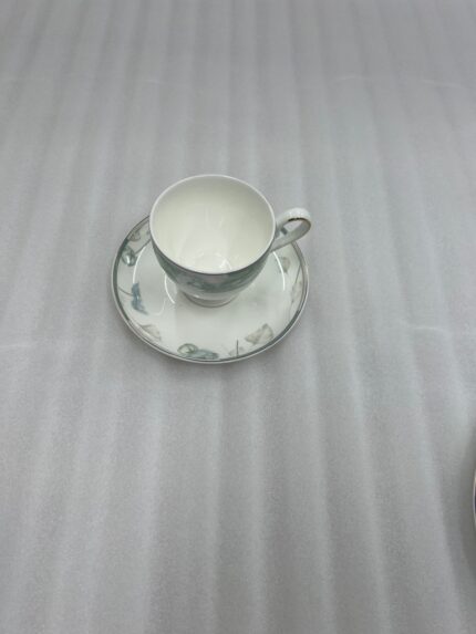 A luxury super bone china tea cup with a floral mint-green band and gold rim, filled with coffee, resting on a matching saucer on a polished wooden table in a classic living room with a fireplace
