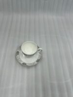 A high-angle shot of a floral-patterned white bone china teacup filled with steaming tea, resting on a matching saucer on a polished wooden table in a luxurious library setting with a green velvet sofa and bookshelves