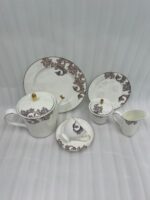 An elegant white and grey floral tea set on a wooden table, featuring a person pouring milk into a cup of tea alongside a plate of toasted rusks.