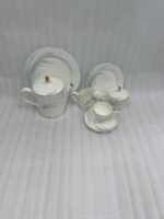 A luxury white Super Bone China tea set with gold rims and a grey feather design, featuring a teapot, creamer, sugar bowl, and cup and saucer on a rustic wooden garden table at sunset