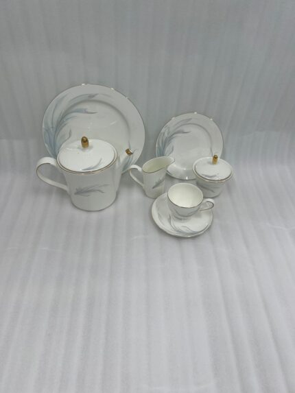 A luxury white Super Bone China tea set with gold rims and a grey feather design, featuring a teapot, creamer, sugar bowl, and cup and saucer on a rustic wooden garden table at sunset
