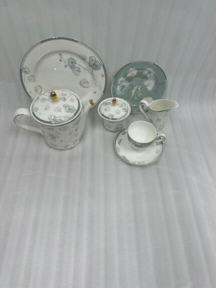 A person pouring milk from a floral bone china creamer into a teacup on a wooden table, surrounded by a matching tea set, teapot, and a plate of sliced cake.