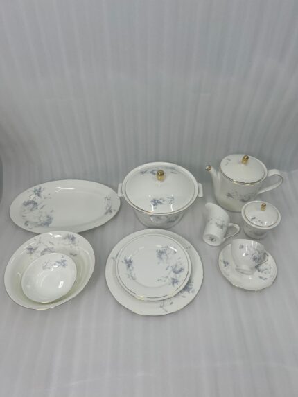 High-angle view of a 61-piece white super bone china dinner set with blue floral patterns and gold rims, elegantly arranged for an 8-person serving on a long mahogany dining table with blue chairs.
