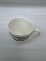 A high-quality white bone china mug filled with steaming milk tea, featuring a gold Greek Key pattern and beaded texture, resting on a rustic wooden garden table