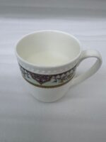 A high-angle close-up of a white bone china mug filled with green tea, featuring an intricate burgundy and gold damask pattern on a mint-green band, resting on a beige linen napkin on a wooden table with books and white rose petals in the background