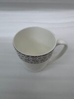 A steaming cup of latte with latte art inside a white bone china mug featuring a gold geometric pattern and quilted texture, resting on a glass saucer with a silver spoon on a wooden table