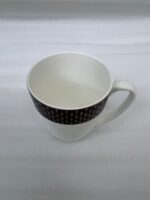 A high-angle shot of a premium white bone china mug with a textured diamond pattern and a dark geometric border, filled with latte art on a wooden table alongside a leather journal and coffee beans