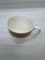 A high-quality bone china coffee mug with a latte inside, featuring a gold woven pattern at the top and an embossed white grid texture on the bottom, set on a dark marble countertop next to coffee beans and a gold spoon