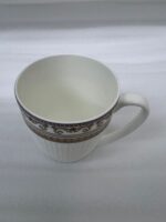 A high-angle shot of a luxury white bone china mug filled with black coffee, featuring an ornate gold Greek key border, placed on a dark wooden table next to a silver spoon in a cozy living room setting