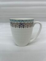 A high-angle close-up of a white bone china mug filled with milk, featuring a gold filigree border and textured lattice design, resting on a rustic blue wooden table in a brightly lit room
