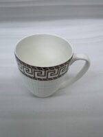 A close-up of a steaming mug of tea with a white and gold Greek Key pattern on a wooden table, part of a 6-piece Bone China mug set