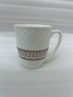 A close-up shot of a white bone china mug with an embossed diamond pattern and a brown Greek key design, filled with latte art, sitting on a granite kitchen countertop next to a silver spoon