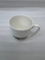 A close-up shot of a white bone china mug filled with tea, resting on a slate coaster on a rainy balcony railing. The mug features an embossed diamond pattern and a decorative floral band at the bottom