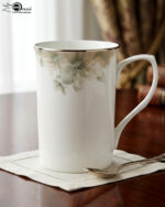A professional product shot labeled 3.jpg showing two elegant bone china mugs with floral patterns and gold rims on a wooden table, set against a classic living room background with an armchair and fireplace
