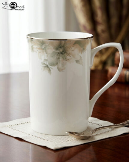A professional product shot labeled 3.jpg showing two elegant bone china mugs with floral patterns and gold rims on a wooden table, set against a classic living room background with an armchair and fireplace