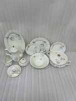 An elegant, white super bone dinner set with a delicate blue rose floral pattern and gold rims, displayed on a beige marble tabletop against a neutral background. The set includes dinner plates, side plates, a large oval serving platter, a covered casserole dish, a teapot, a sugar bowl, a creamer pitcher, a teacup with a saucer, and serving bowls