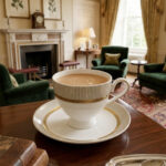 A luxury super bone china tea cup and saucer with gold pinstripe and laurel wreath patterns, filled with milk tea on a wooden table in a classic vintage living room setting with a fireplace and green armchairs