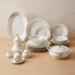 An elegant white super bone china dinner set with gold rims and a delicate grey leaf pattern, displayed on a wooden table