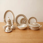 An elegant super bone china dinnerware set featuring a fine beige and gold-lined sunburst pattern, arranged neatly on a wooden table. The collection includes dinner plates, an oval platter, a tureen with a lid, soup bowls, a teapot, a creamer, a sugar bowl, and a teacup with a saucer