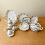 An elegant white and grey floral patterned super bone china dinner set arranged on a wooden table, including plates, bowls, a teapot, a casserole dish, and teacups.