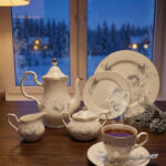 A luxury white super bone china tea set with grey floral patterns and gold rims on a wooden table, featuring a teapot, teacup, plates, and a steaming cup of tea against a snowy window BG.