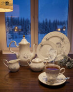A luxury white super bone china tea set with grey floral patterns and gold rims on a wooden table, featuring a teapot, teacup, plates, and a steaming cup of tea against a snowy window BG.