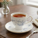 A steaming cup of black tea in a white bone china cup featuring blue floral patterns and a gold rim, resting on a matching saucer on a dark wooden table