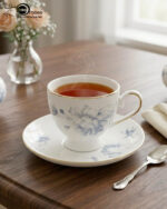 A steaming cup of black tea in a white bone china cup featuring blue floral patterns and a gold rim, resting on a matching saucer on a dark wooden table