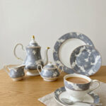 A premium grey and white floral pattern super bone china tea set featuring a teapot with gold accents, a creamer, a sugar bowl, plates, and a filled teacup with a saucer on a wooden table.