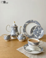 A premium grey and white floral pattern super bone china tea set featuring a teapot with gold accents, a creamer, a sugar bowl, plates, and a filled teacup with a saucer on a wooden table.