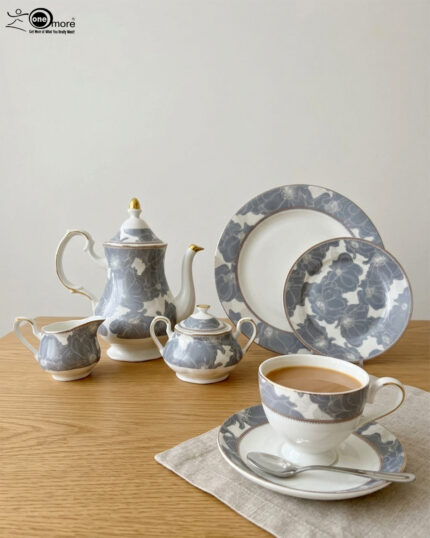 A premium grey and white floral pattern super bone china tea set featuring a teapot with gold accents, a creamer, a sugar bowl, plates, and a filled teacup with a saucer on a wooden table.