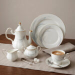 Luxury super bone china tea set featuring a teapot, teacup, saucer, creamer, and sugar bowl with fine gold trim and soft grey feather patterns on a wooden table.