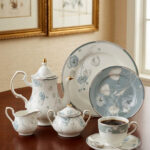 A premium super bone china tea set featuring a classic teapot, sugar bowl, creamer, teacup with saucer, and matching plates, all adorned with an elegant light blue and grey floral pattern on a wooden table.