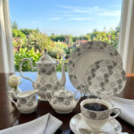 A premium bone china tea and coffee set with a grey floral pattern and silver rims on a dark wooden table. The collection includes a teapot, creamer pitcher, sugar bowl, plates, and a steaming cup of black coffee on a saucer with a silver spoon, set against a bright window showing a lush garden background
