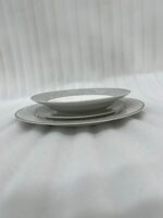 A stacked 18-piece elegant super bone china plate set on a dark wooden dining table. The set includes quarter plates on top, deep plates in the middle, and large rice plates at the bottom, all featuring a light grey floral pattern border and gold trim