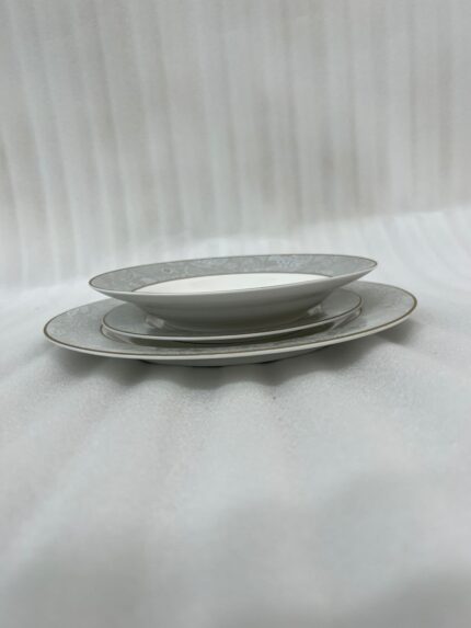 A stacked 18-piece elegant super bone china plate set on a dark wooden dining table. The set includes quarter plates on top, deep plates in the middle, and large rice plates at the bottom, all featuring a light grey floral pattern border and gold trim