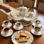 An elegant white and grey floral tea set on a wooden table, featuring a person pouring milk into a cup of tea alongside a plate of toasted rusks.