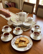 An elegant white and grey floral tea set on a wooden table, featuring a person pouring milk into a cup of tea alongside a plate of toasted rusks.