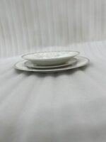 A stacked 18-piece elegant white super bone china dinner plate set featuring a delicate grey floral pattern on the rims, neatly arranged on a grey fabric runner over a wooden dining table with crystal glasses and a silver pitcher in the background.