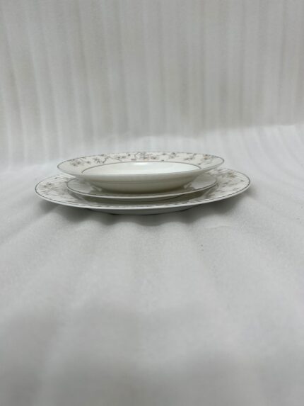 A stacked 18-piece premium super bone china dinnerware set with elegant floral and striped rim borders, neatly arranged on a dark wooden dining table next to a crystal glass and fabric napkin