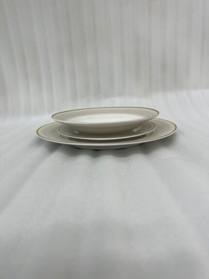 A neatly stacked 18-piece super bone china dinner plate set with elegant gold borders, arranged on a beige placemat next to a gold fork, a gold knife, and a crystal wine glass on a wooden table.