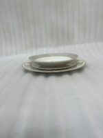 A stacked 18-piece super bone china dinner plate set featuring a white porcelain body with an elegant gold geometric border pattern, neatly arranged on a white damask table runner over a dark wooden dining table next to a rolled napkin with a gold ring.