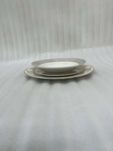 A stacked 18-piece super bone china dinner plate set featuring a white porcelain body with an elegant gold geometric border pattern, neatly arranged on a white damask table runner over a dark wooden dining table next to a rolled napkin with a gold ring.