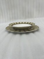 A stacked 18-piece super bone china plate set neatly arranged on a beige linen placemat over a wooden dining table. The ceramic dinnerware set features 6 quarter plates stacked on top, 6 deep soup plates in the middle, and 6 large rice plates at the bottom. Each plate showcases an elegant radiating striped pattern rim in olive green, cream, and taupe tones with fine gold rim detailing. A blur background shows a window and a small flower vase